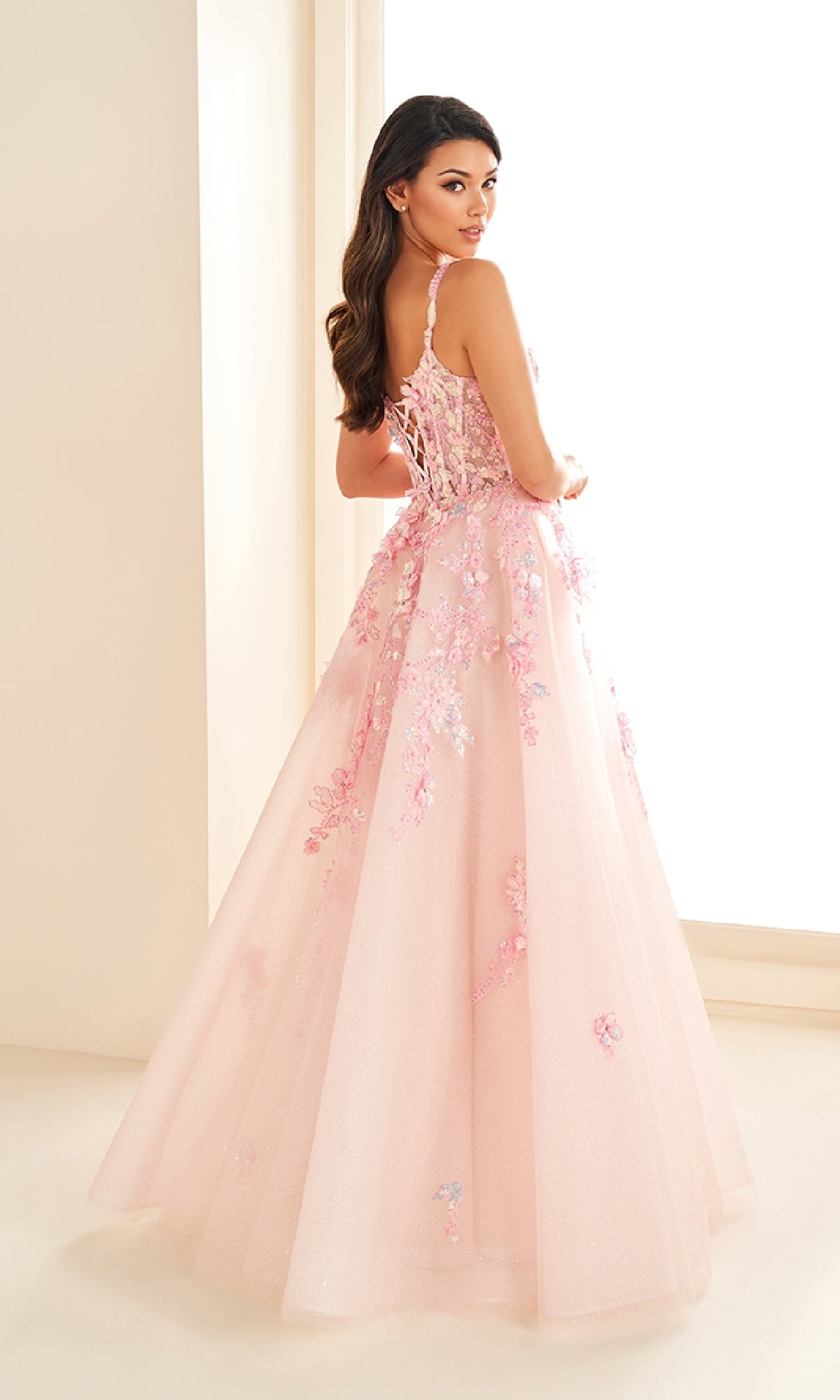 A woman in the Ellie Wilde EW36213 long prom dress—light pink glitter tulle with floral lace appliqués and a lace-up back—stands in a softly lit room, facing slightly away from the camera.